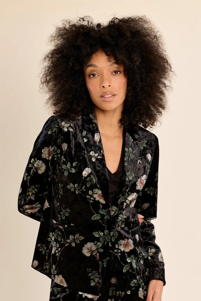 A model with voluminous dark curly hair wearing a black velvet pajama set with a floral print, featuring a long-sleeve top and matching pants, against a solid beige background.