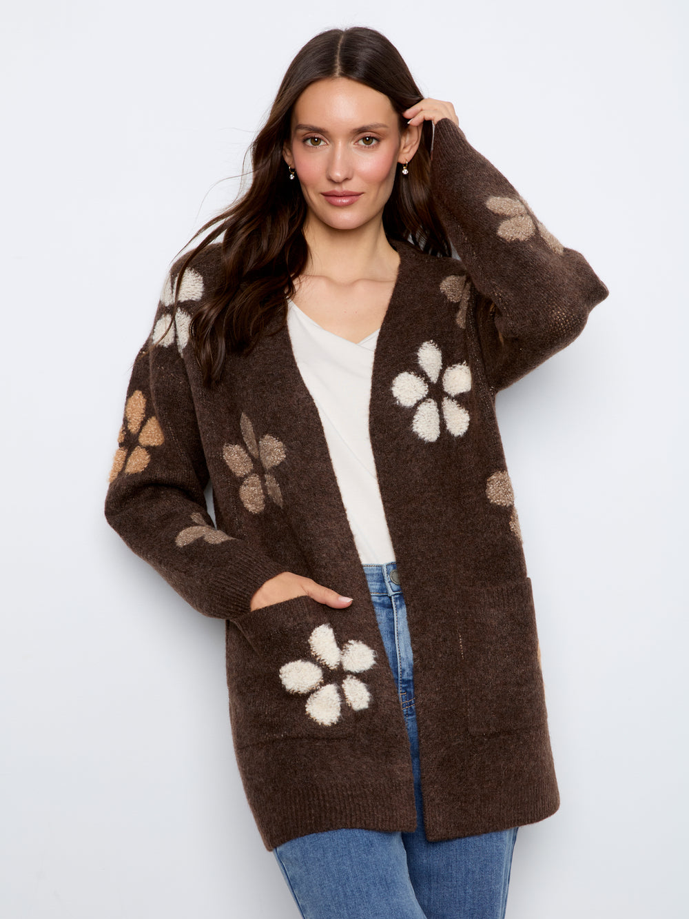 A woman modeling a brown open-front cardigan with large cream and beige floral knit patterns, worn over a white top and blue jeans against a light gray studio background.