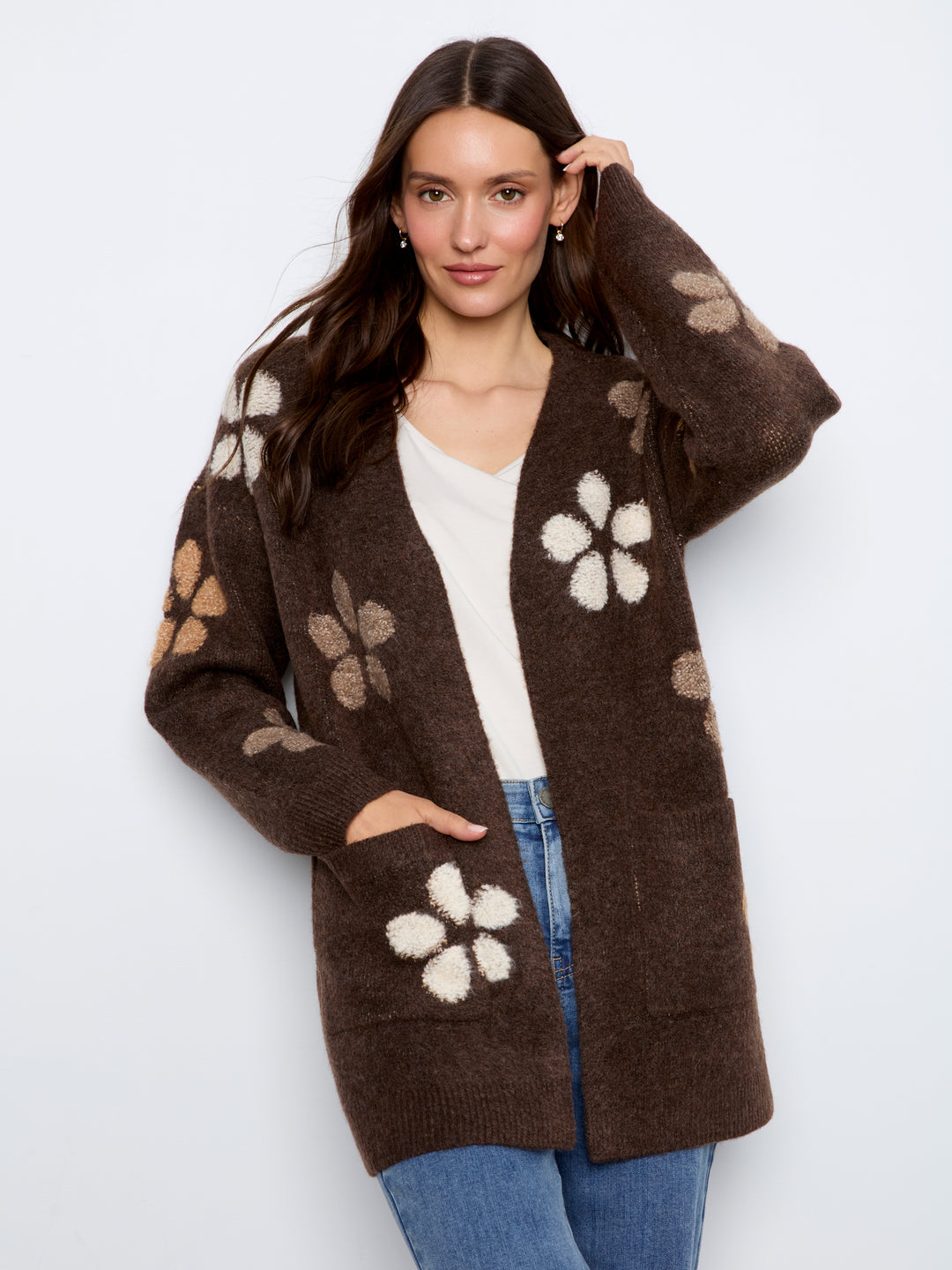 A woman modeling a brown open-front cardigan with large cream and beige floral knit patterns, worn over a white top and blue jeans against a light gray studio background.