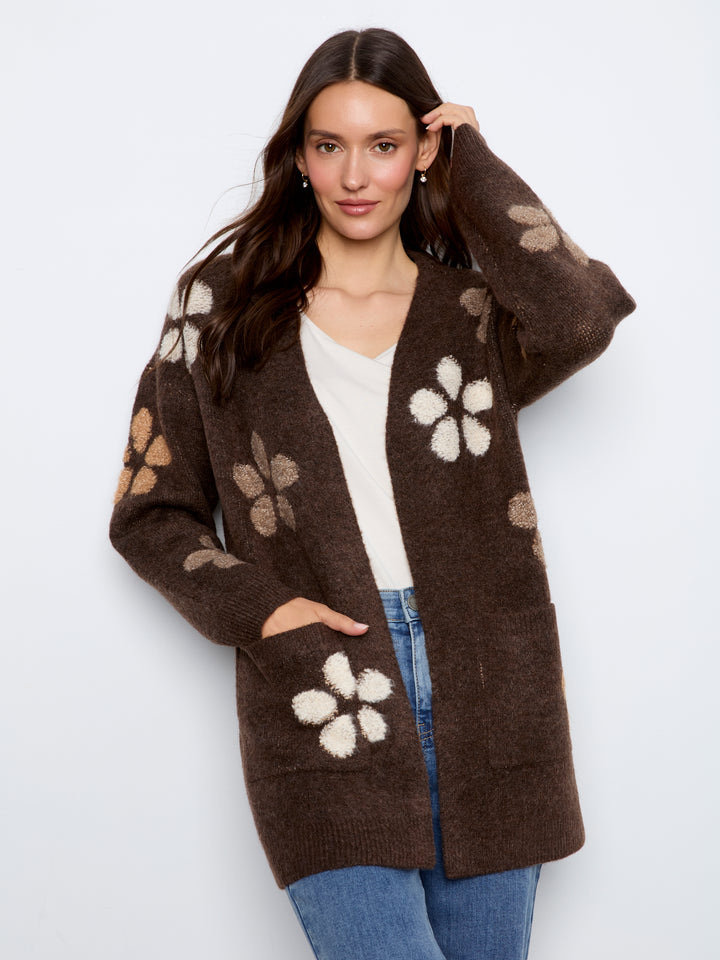 A woman modeling a brown open-front cardigan with large cream and beige floral knit patterns, worn over a white top and blue jeans against a light gray studio background.