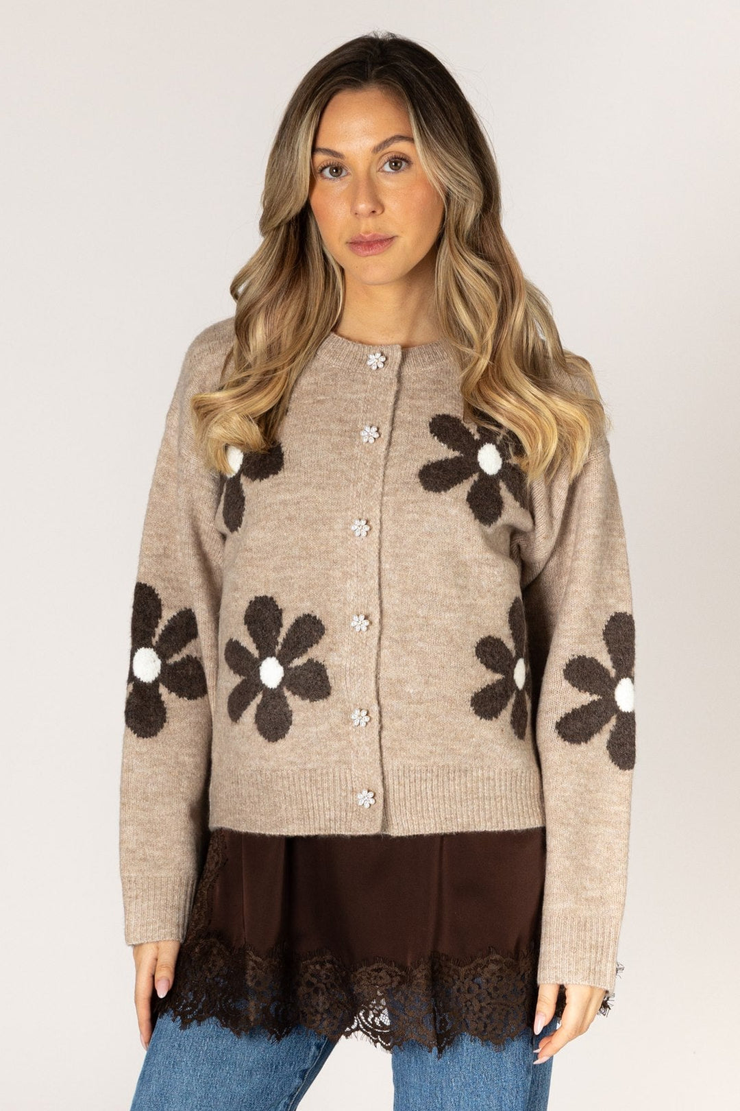 A woman modeling a beige cardigan with large dark brown flower motifs and white centers, paired with a dark brown top and blue jeans, against a plain light gray background.
