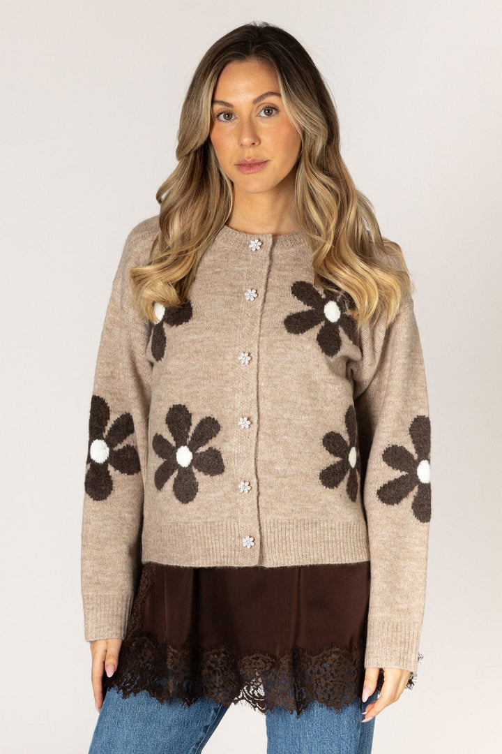 A woman modeling a beige cardigan with large dark brown flower motifs and white centers, paired with a dark brown top and blue jeans, against a plain light gray background.