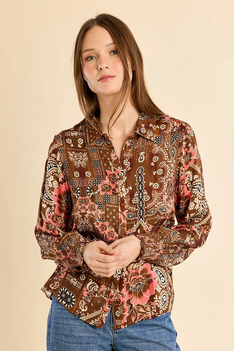 A woman modeling a long-sleeve button-up blouse with an intricate brown, pink, and beige paisley and floral pattern, paired with blue jeans against a solid beige background.