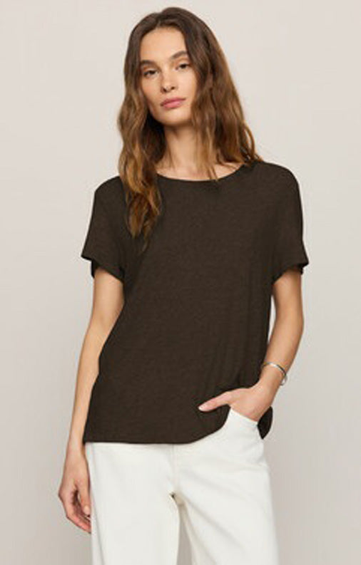 A woman modeling a dark brown short-sleeve knit top with a wide boat neckline, paired with white pants, against a light beige studio background.