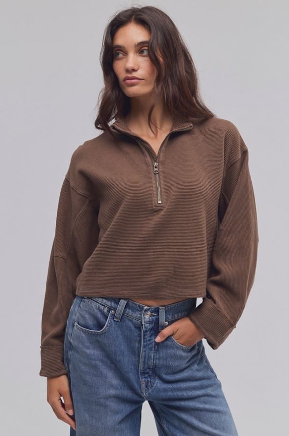 A woman wearing a brown ribbed quarter-zip pullover sweater and blue jeans, standing against a solid gray studio background.