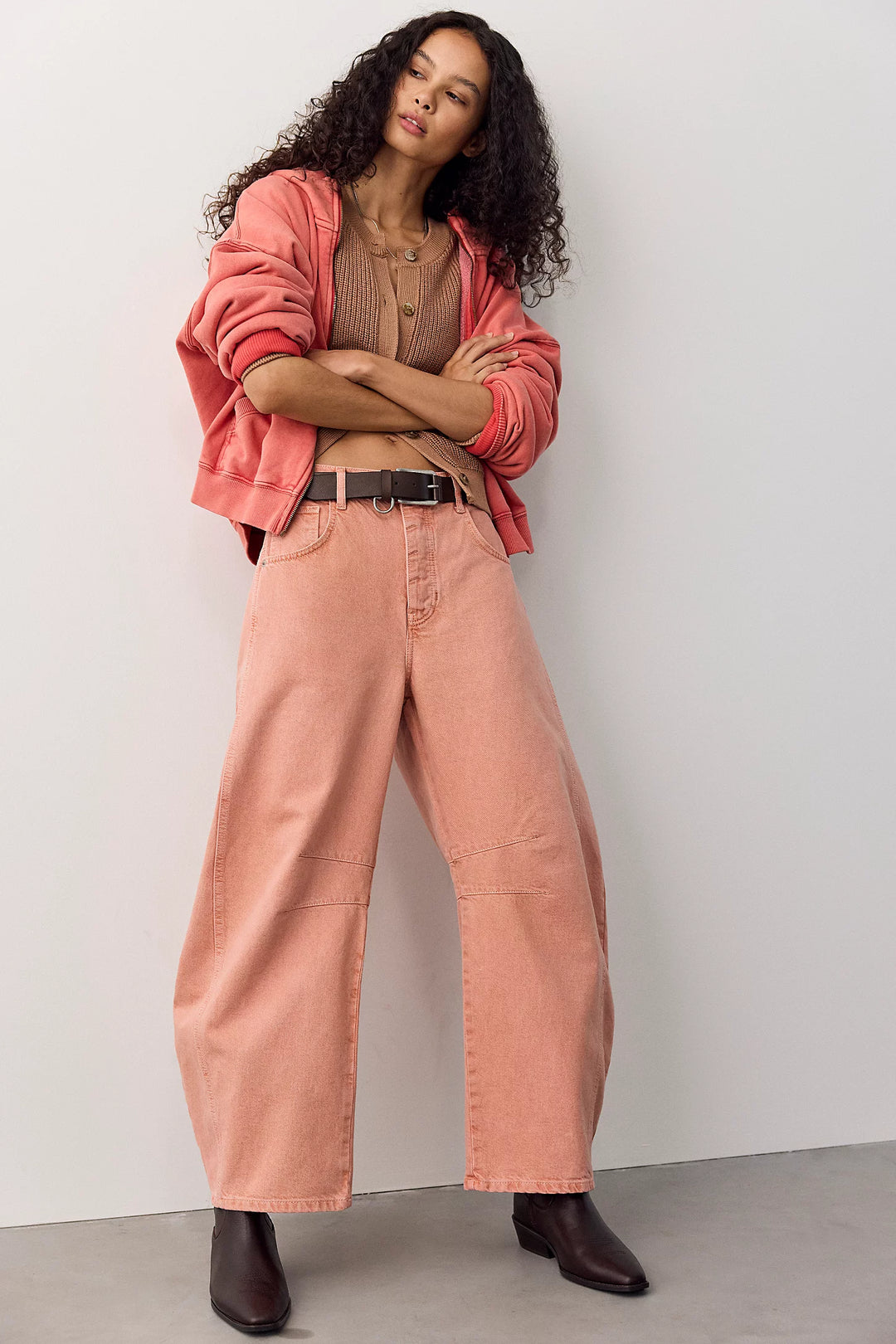 A model stands against a light gray wall wearing wide-leg dusty pink jeans, a brown top, a black belt, a coral pink cropped jacket, and dark brown ankle boots.