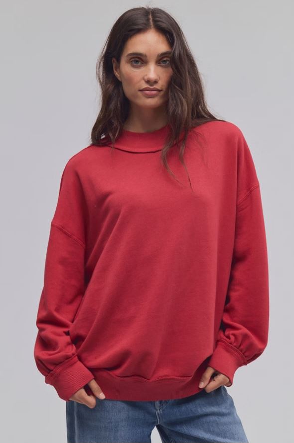 A woman wearing an oversized red crewneck sweatshirt and blue jeans, standing against a solid light gray studio background.