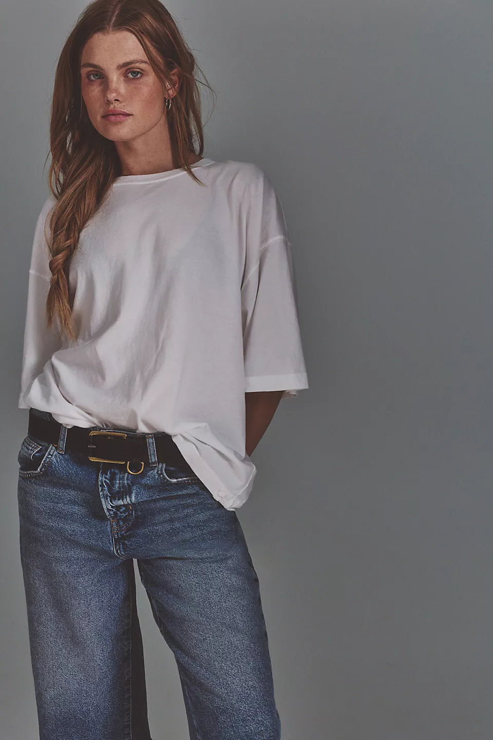 A woman wearing an oversized white short-sleeve t-shirt tucked into blue jeans with a black belt, standing against a solid gray studio background.
