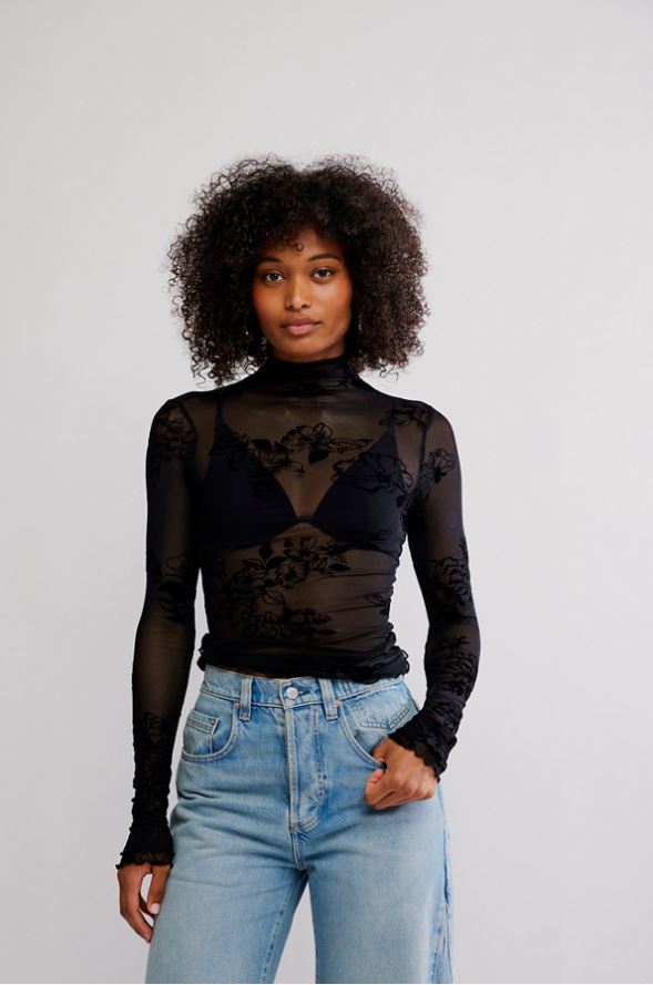 A woman with curly dark hair wearing a sheer black long-sleeve mock-neck top with floral lace applique over a black bralette, paired with light-wash blue jeans, against a plain light gray studio background.