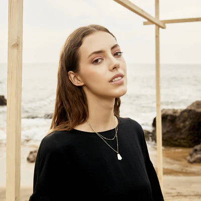 Woman wearing a black top and layered necklaces standing on a beach with wooden structures.