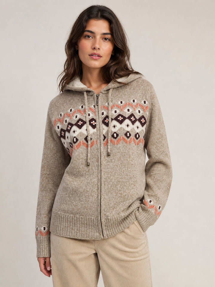 A woman wearing a beige zip-up hooded sweater with a colorful geometric pattern across the chest and cuffs, paired with beige pants, against a plain light gray studio background.