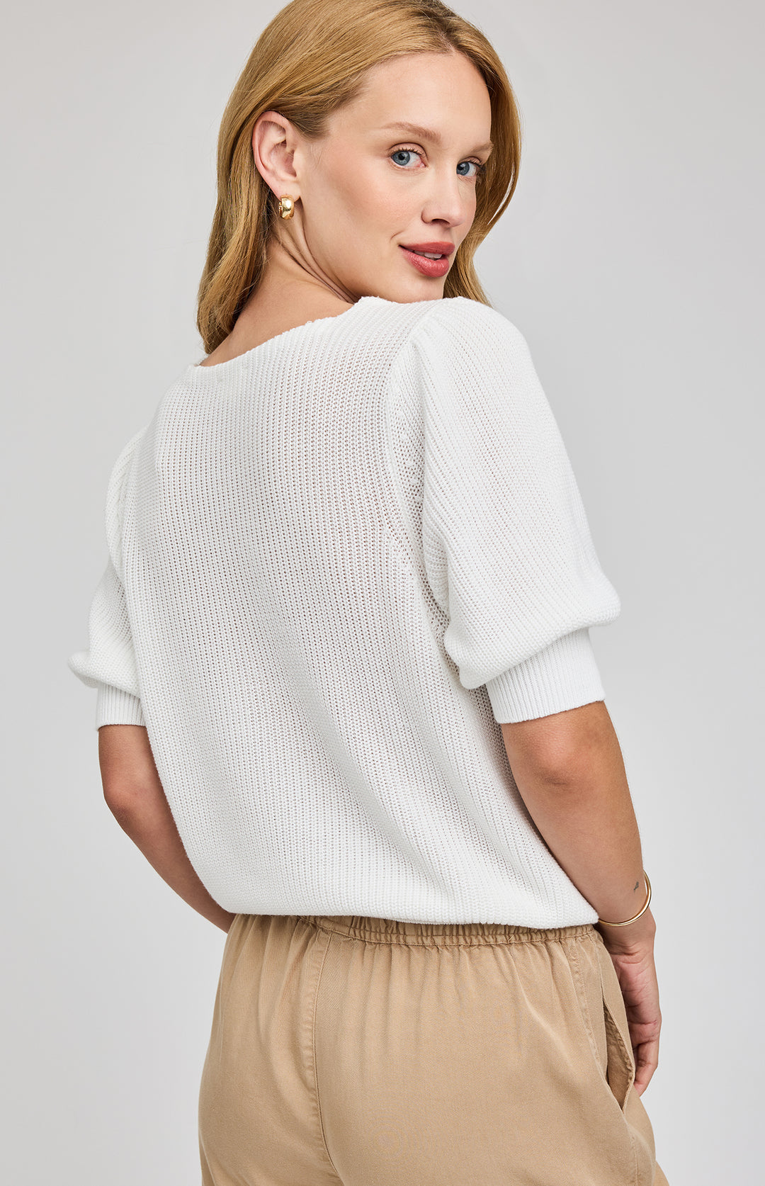 Woman wearing a white sweater and beige pants against a plain background