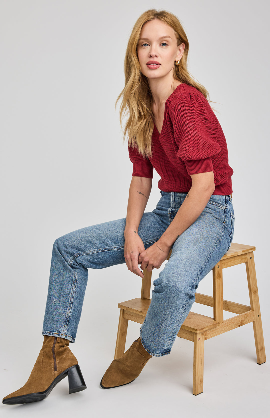 Woman sitting on a wooden stool wearing a red top and blue jeans against a white background