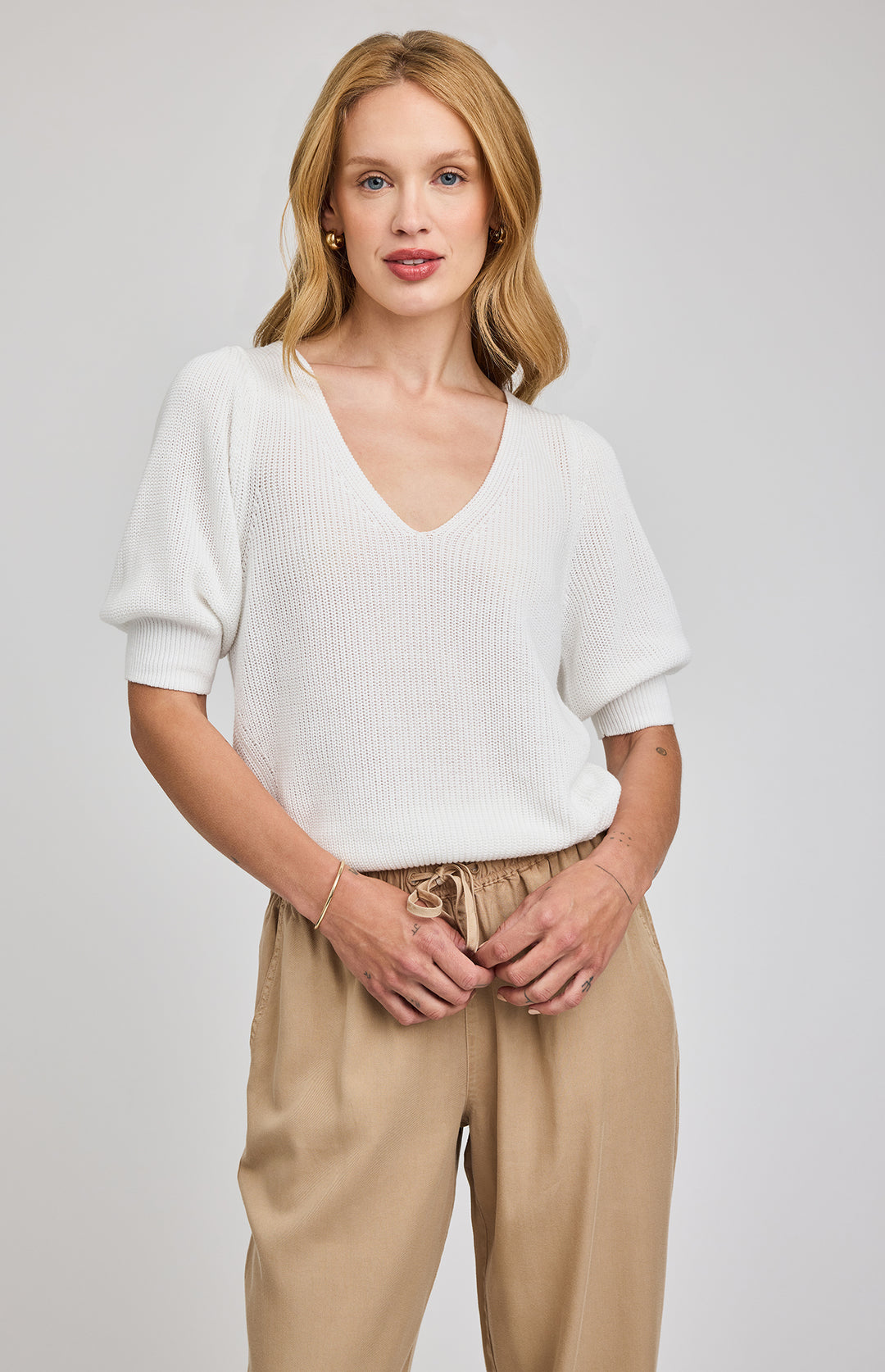 Woman wearing a white sweater and beige pants against a plain background