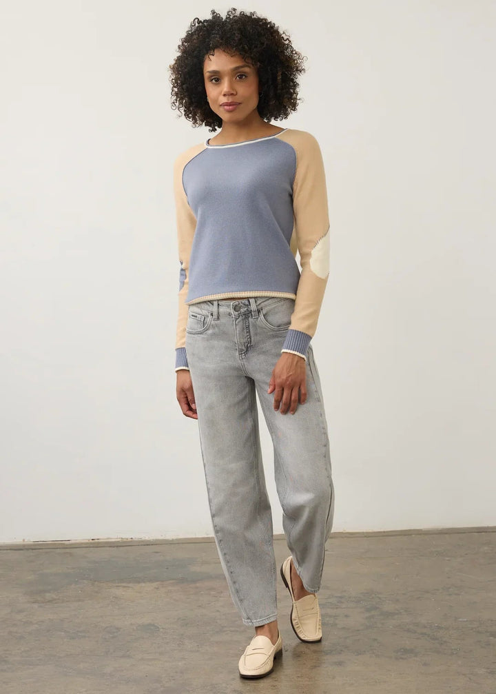 A model stands in a minimalist studio setting wearing a color-blocked raglan long-sleeve top with a dusty blue body and beige sleeves, paired with light-wash jeans and beige loafers.