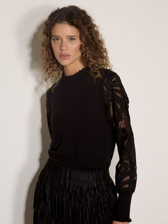 A woman with curly brown hair wearing a black long-sleeve knit top with sheer lace sleeves and a black pleated skirt, posing against a plain off-white wall.
