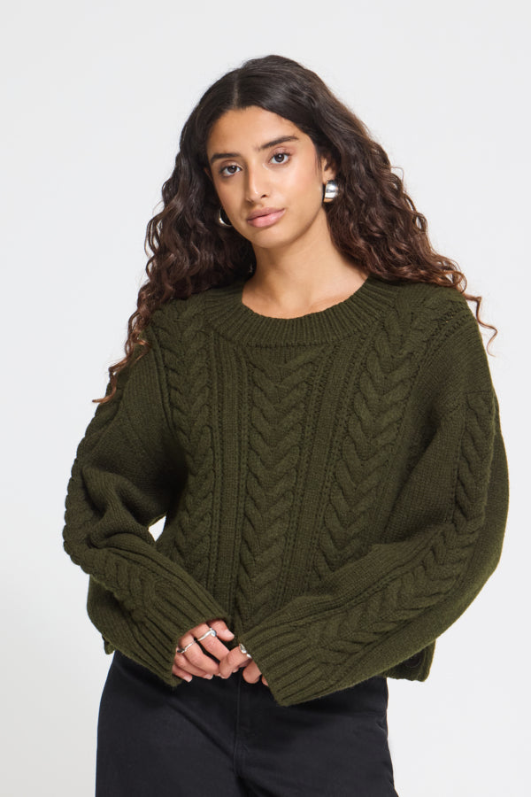 A woman with long, wavy dark brown hair wearing an olive green cable-knit sweater and black pants, posing against a light gray studio background.