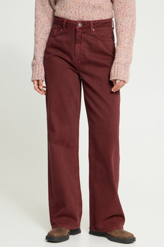A person wearing a pair of wide-leg straight denim jeans in a deep burgundy color, paired with a light pink speckled knit sweater and brown suede boots, shown from the waist down on a white studio background.