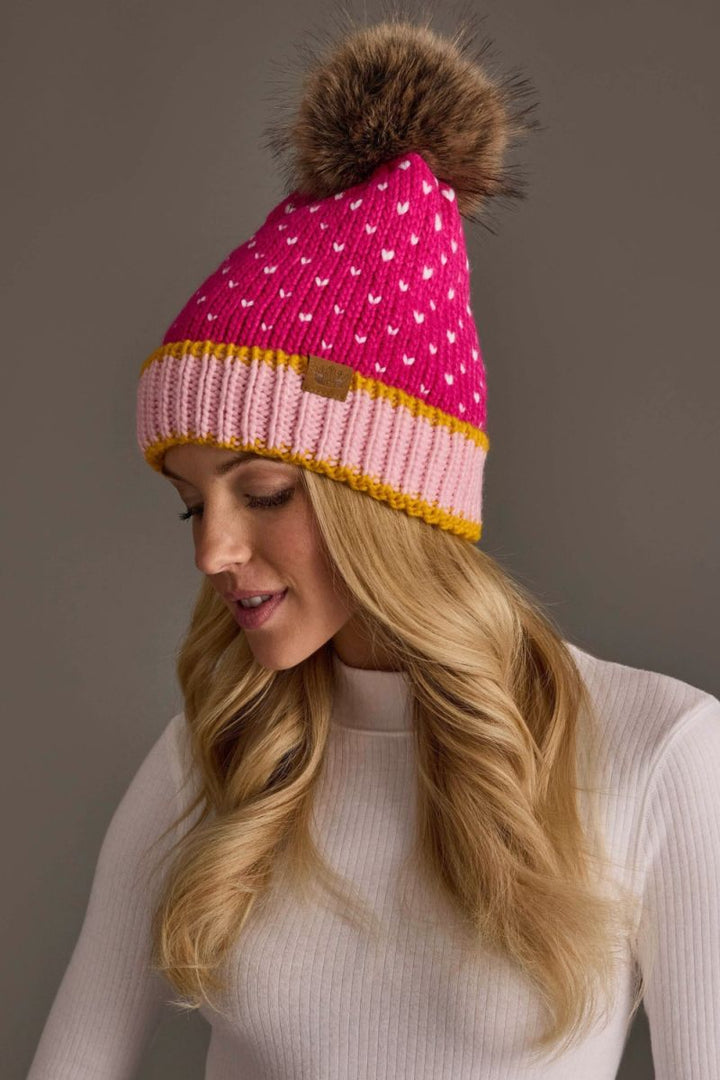 A woman wearing a bright pink knit beanie with white heart patterns, a yellow stripe, and a brown faux-fur pom-pom. She is wearing a white ribbed turtleneck sweater against a solid gray studio background.
