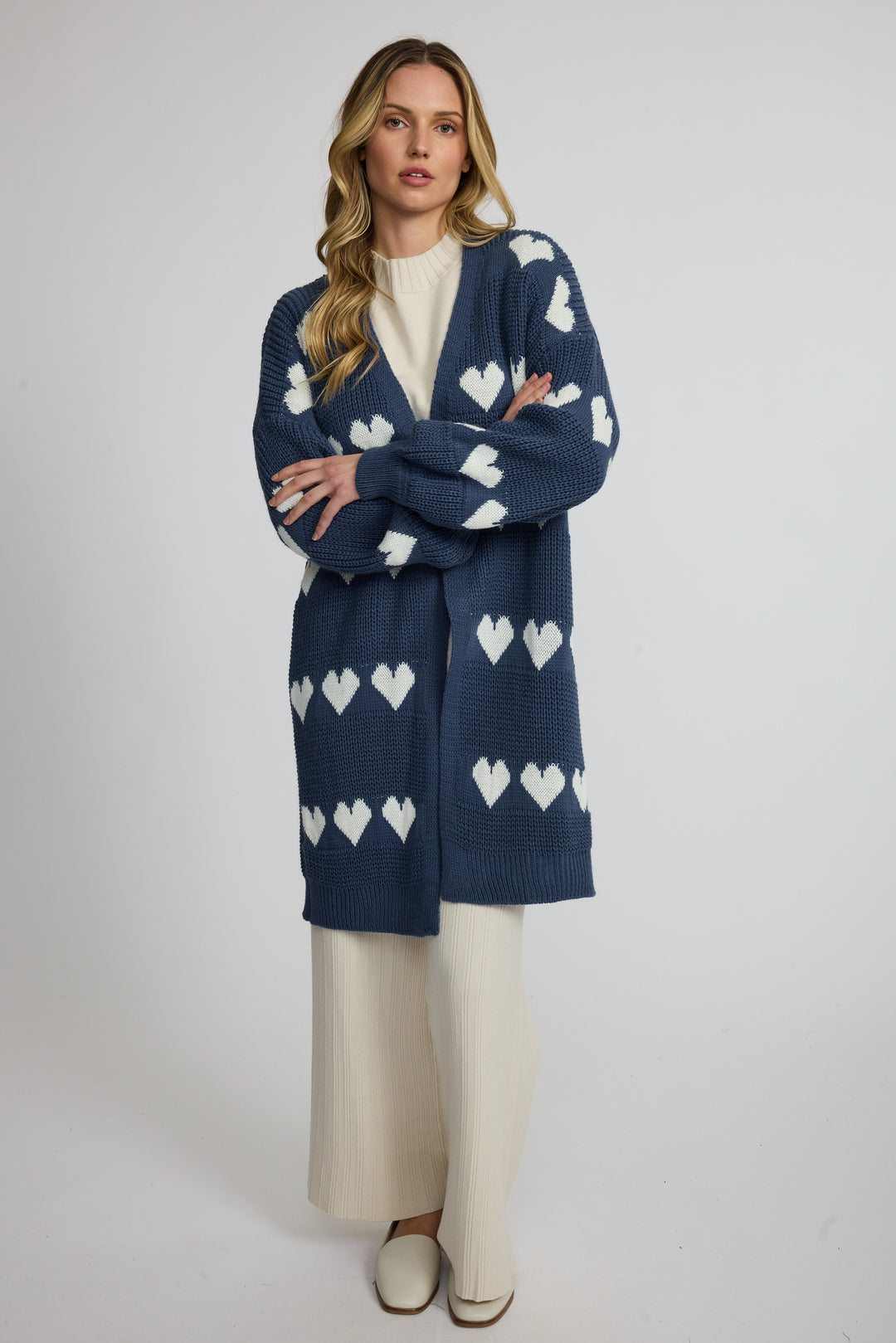A woman modeling a navy blue knit cardigan with white heart patterns, worn over a cream-colored top and wide-leg cream pants against a light gray studio background.