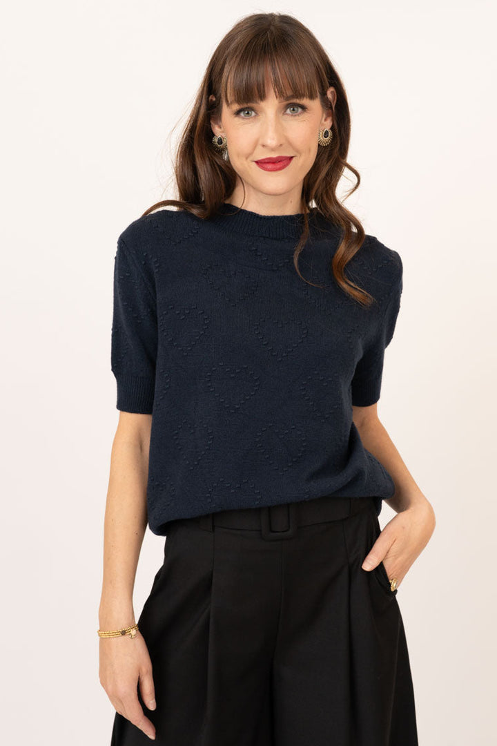 A woman modeling a short-sleeve textured knit top in dark navy, paired with black wide-leg trousers, against a plain off-white studio background.