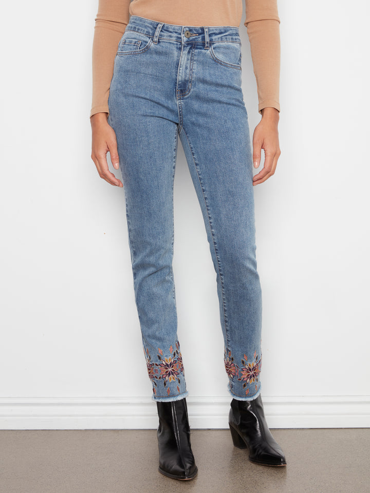 A person wearing light blue high-waisted skinny jeans with floral embroidery near the cuffs, paired with a beige long-sleeve top and black ankle boots, standing against a white wall.