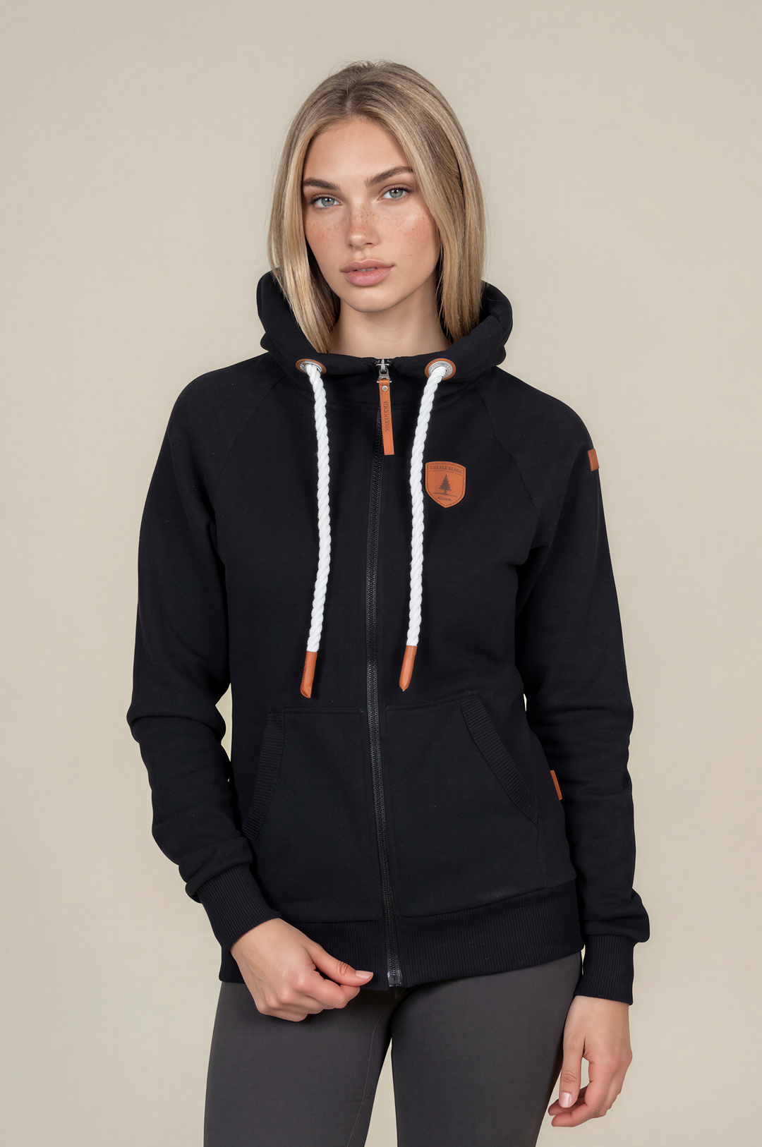 A woman modeling a black zip-up hoodie with white braided drawstrings and orange accents, paired with dark gray leggings, against a solid beige background.