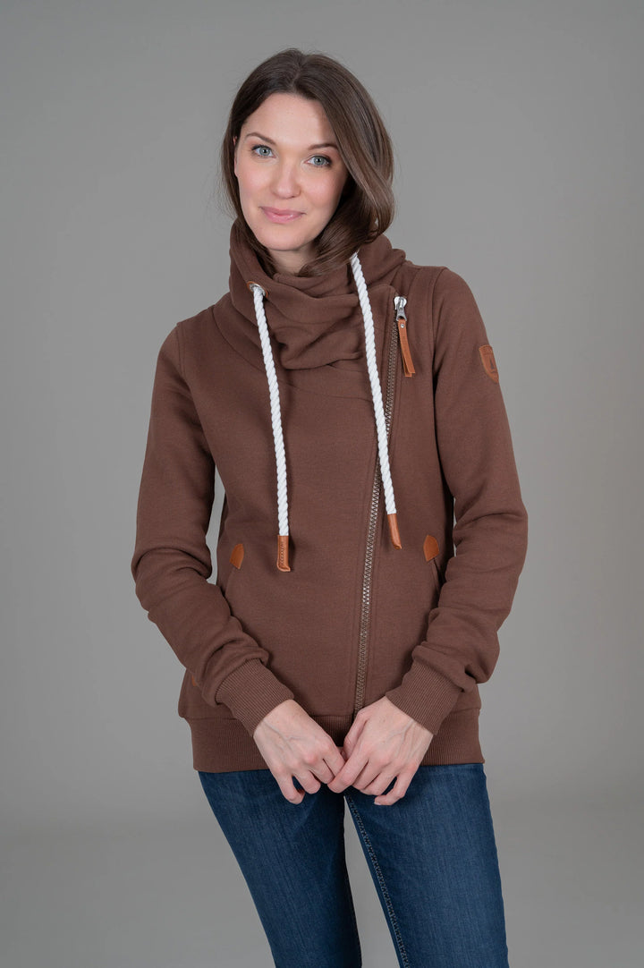 A woman modeling a brown zip-up hoodie with a high cowl neck, white braided drawstrings, and brown leather-like accents, paired with blue jeans against a solid gray studio background.
