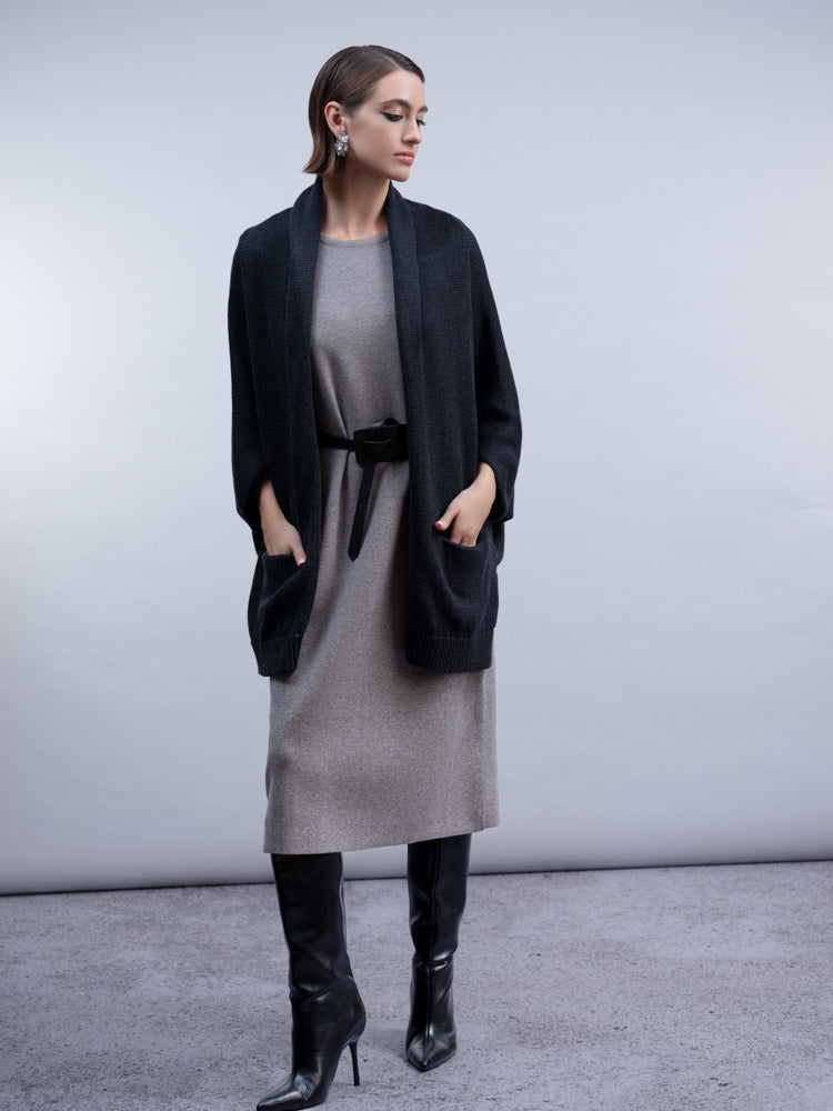 A woman models a black open-front cardigan over a gray knit dress with a black belt, paired with black knee-high boots, against a neutral gray studio background.