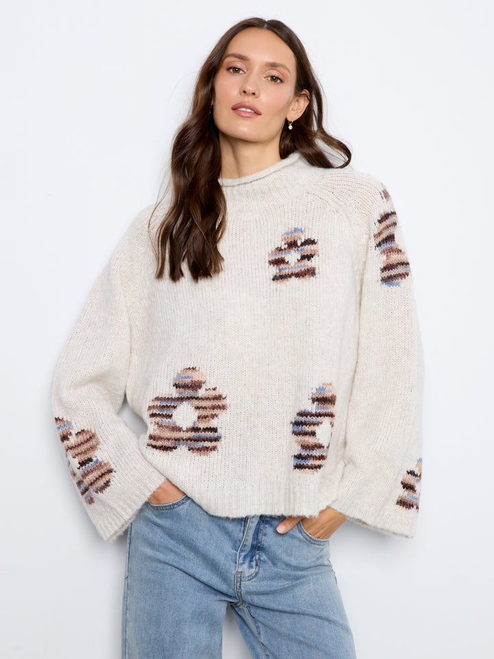 A woman wearing a cream-colored knit sweater with brown and blue striped knit patterns, paired with light blue jeans, against a plain white background.