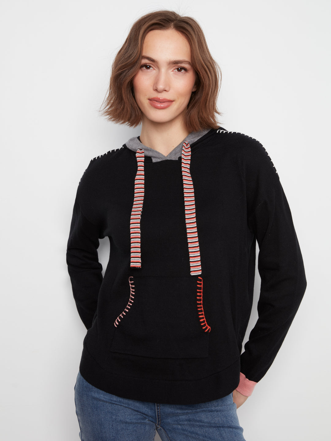 A woman wearing a black pullover hoodie with pink and orange striped drawstrings and decorative stitching on the shoulders and front pockets, paired with blue jeans, against a plain light gray background.