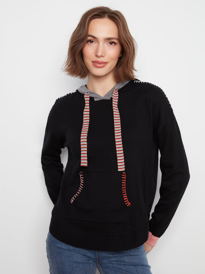 A woman wearing a black pullover hoodie with pink and orange striped drawstrings and decorative stitching on the shoulders and front pockets, paired with blue jeans, against a plain light gray background.