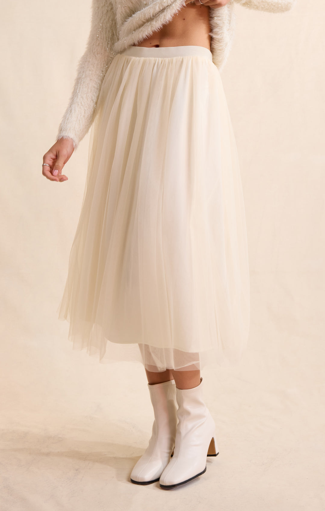 A model wearing a cream-colored tulle midi skirt, styled with a fuzzy knit sweater and white ankle boots against a warm beige background.
