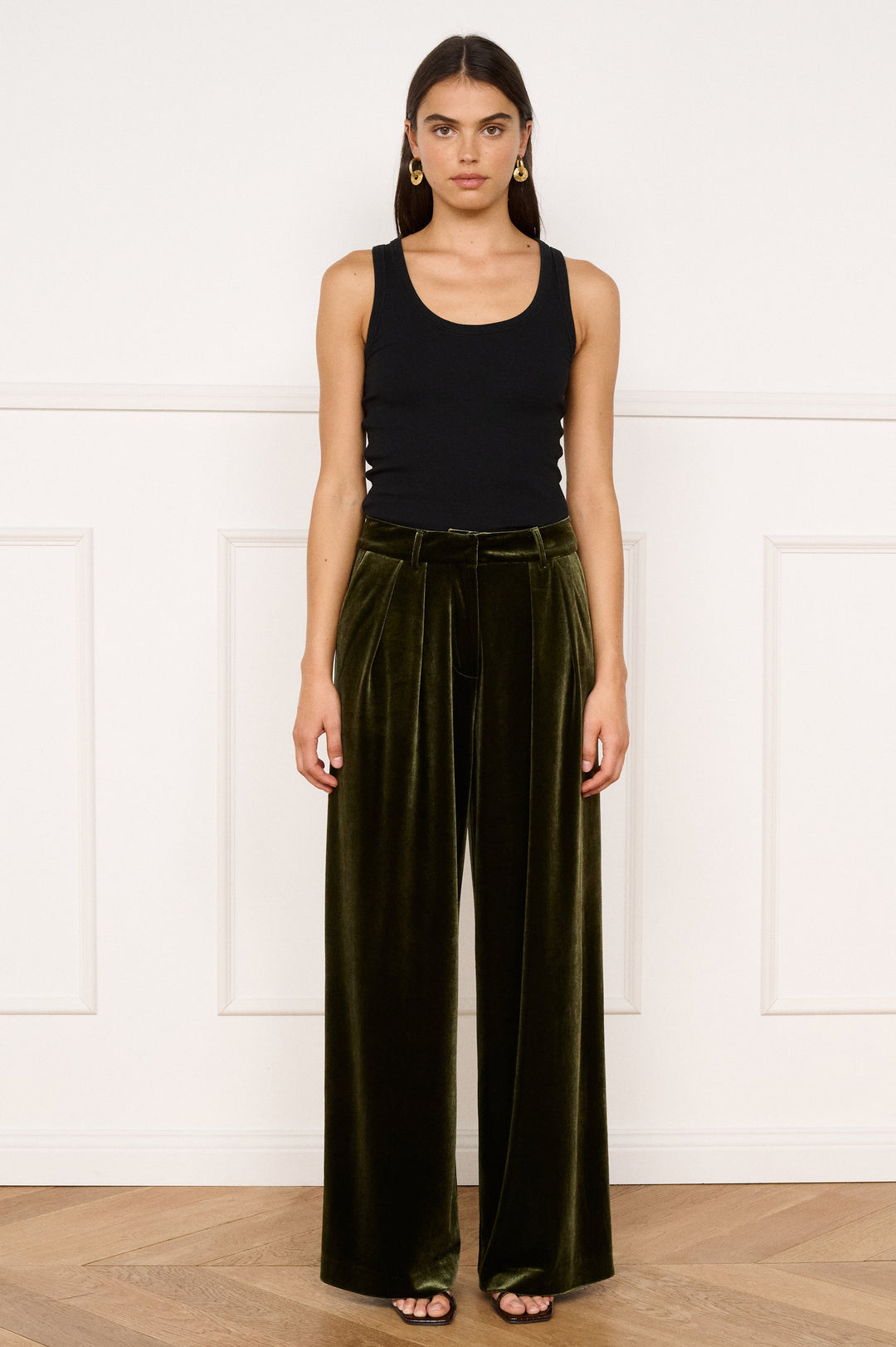 Woman wearing a black tank top and green velvet pants against a white wall.
