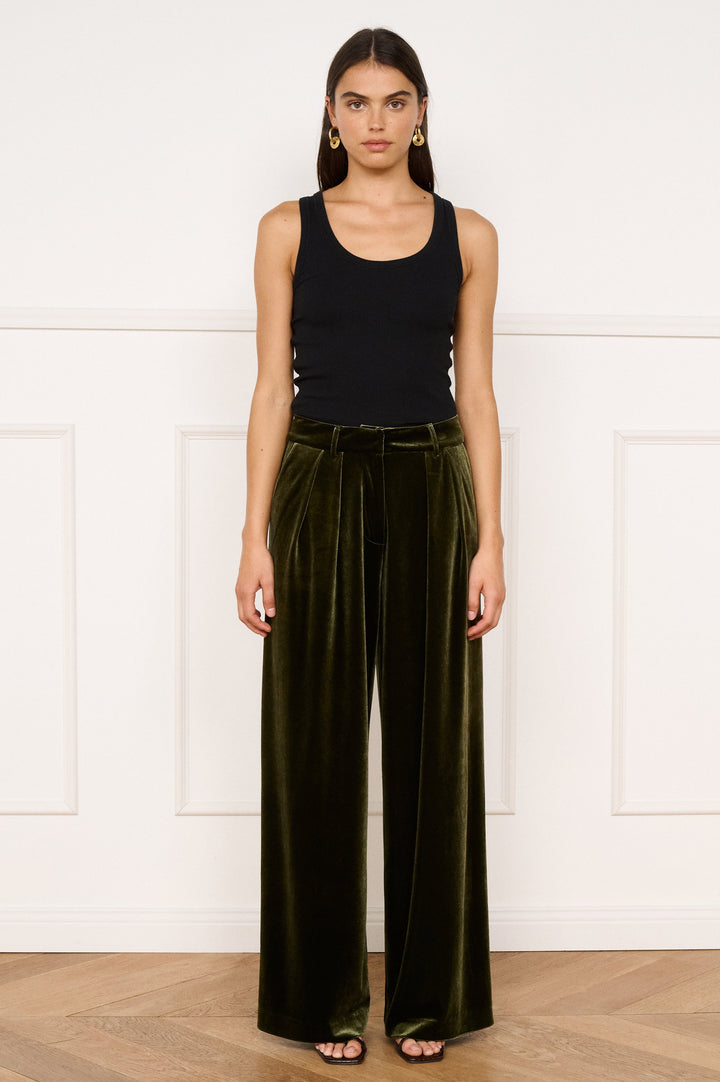 Woman wearing a black tank top and green velvet pants against a white wall.