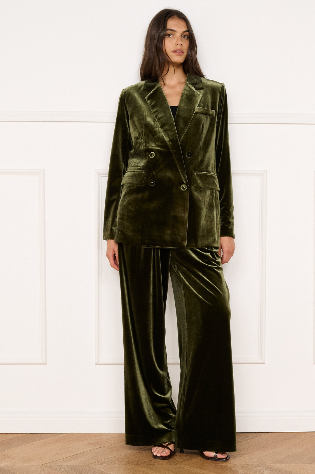 Woman wearing a green velvet suit standing in a room with wooden flooring and white walls.