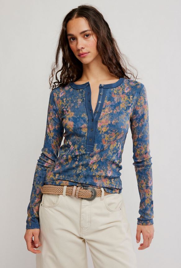 A woman modeling a long-sleeve Henley-style top with a blue floral print, paired with cream pants and a woven belt, against a plain light gray studio background.