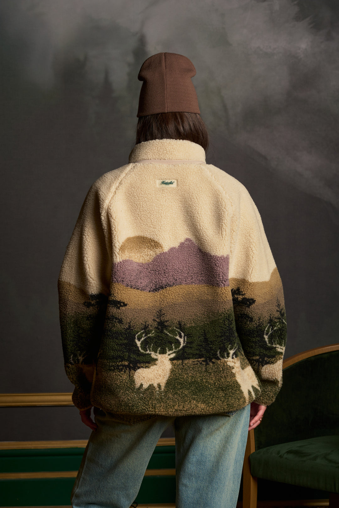 A person viewed from behind wearing a fleece jacket with a scenic mountain and deer pattern, paired with a brown beanie and blue jeans, set against a dark textured studio background.
