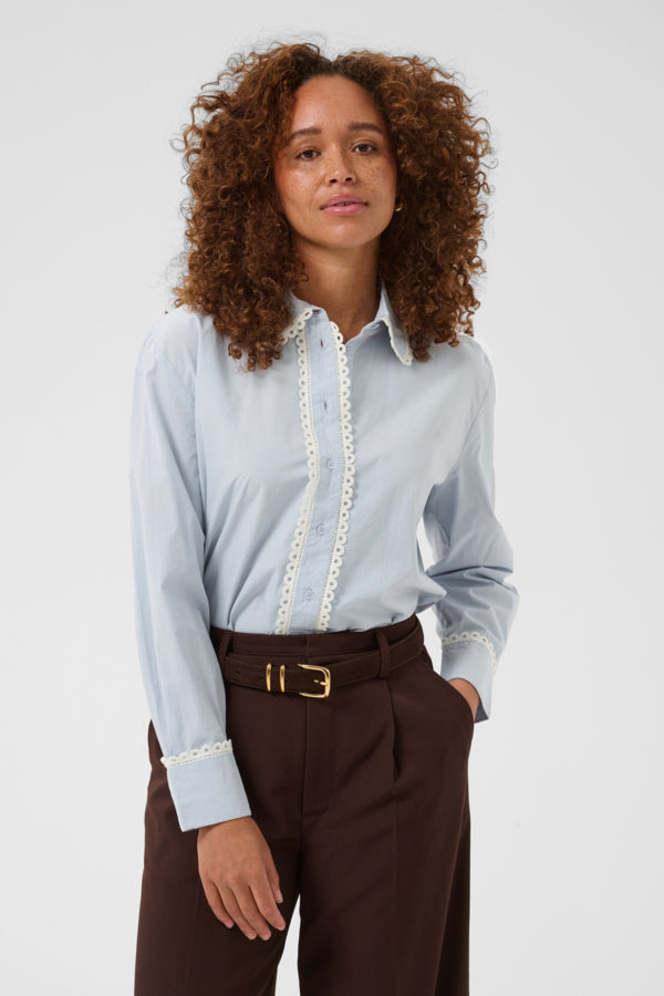 Woman wearing a light blue shirt with lace details and brown pants on a white background