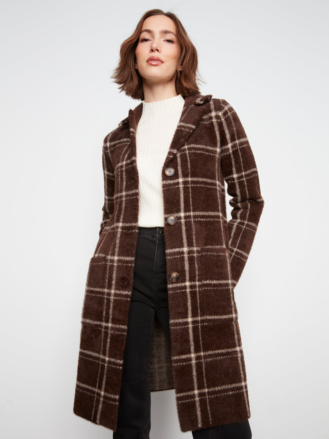 A woman wearing a brown and beige plaid coat over a white top and black pants, photographed against a plain light gray studio background.