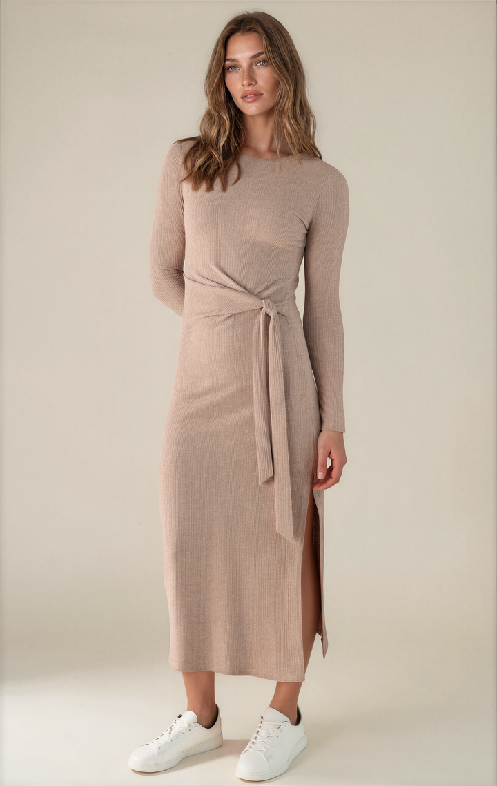 A woman modeling a long-sleeve ribbed knit midi dress in a light taupe-beige color, featuring a wrap-style waist with a tie belt and a side slit. She is wearing white sneakers against a plain, warm off-white studio background.
