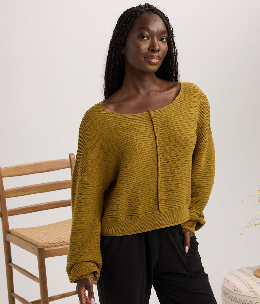 A woman wearing a ribbed mustard yellow long-sleeve top and black pants, standing in a studio setting next to a wooden chair.