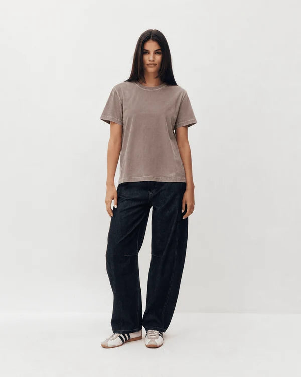 A woman modeling a short-sleeve crewneck t-shirt in a muted taupe-brown color, paired with dark wide-leg jeans and white slide sandals, against a plain white studio background.