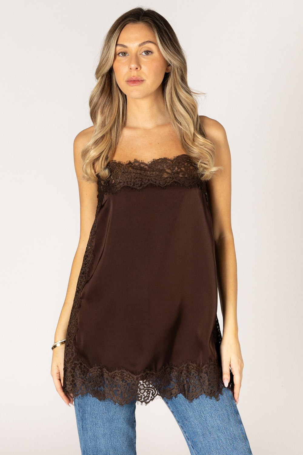 A woman modeling a dark brown satin camisole top with lace trim, paired with blue jeans, against a plain light gray background.