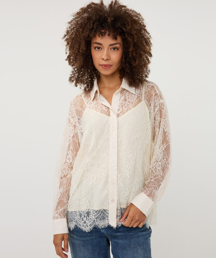 A woman with curly brown hair wearing a cream-colored sheer lace button-up shirt over a matching camisole and blue jeans, photographed against a plain light gray studio background.