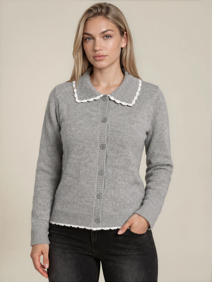 A woman modeling a light gray knit cardigan sweater with a white scalloped Peter Pan collar and matching scalloped hem, paired with black jeans against a solid beige background.