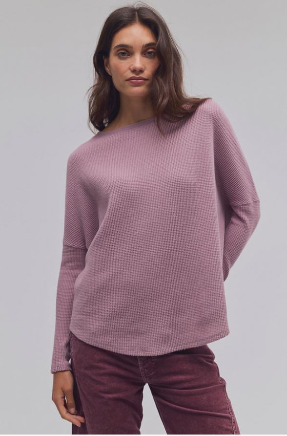 A woman wearing a dusty mauve waffle-knit long-sleeve sweater and burgundy corduroy pants, photographed against a neutral light gray studio background.