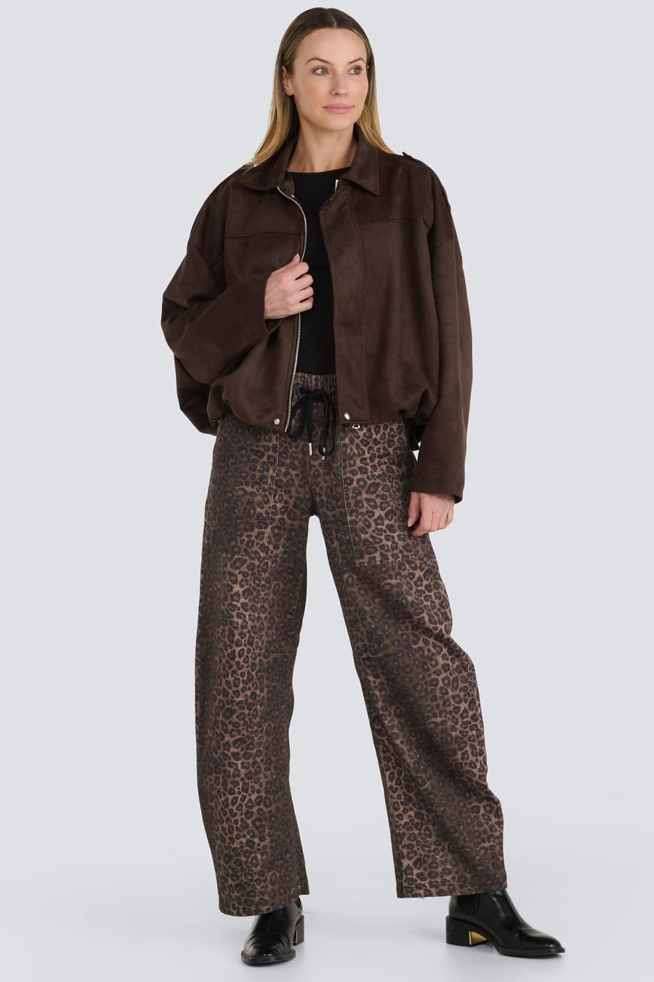 A model stands against a light gray studio background wearing a dark brown suede jacket, wide-leg brown leopard-print trousers, a black top, and black boots.