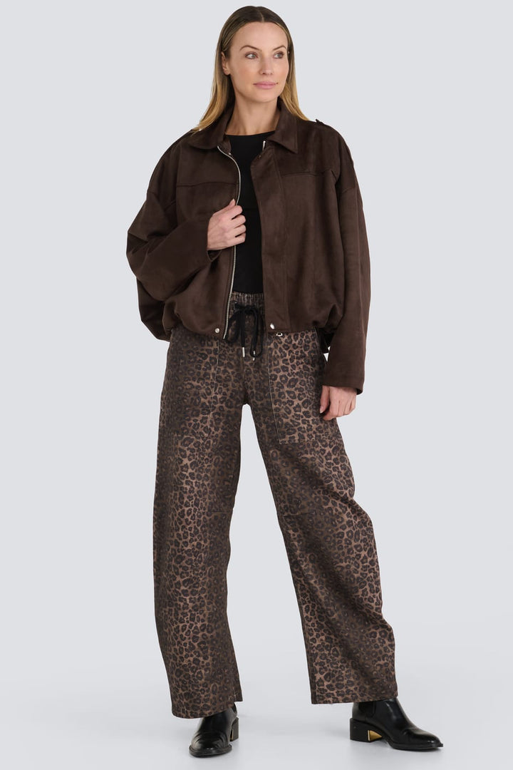 A model stands against a light gray studio background wearing a dark brown suede jacket, wide-leg brown leopard-print trousers, a black top, and black boots.