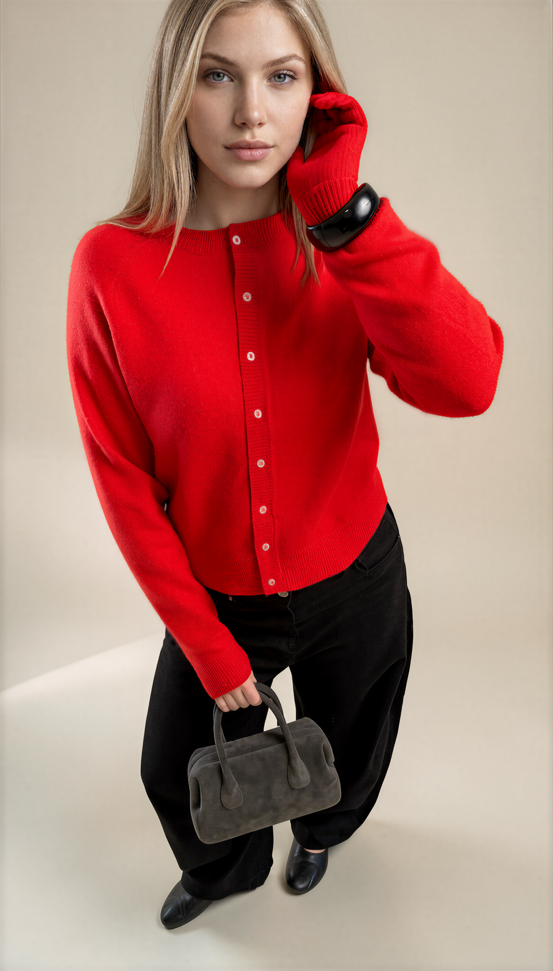 A woman models a bright red knit cardigan sweater with white buttons, paired with black pants and holding a small gray handbag against a warm beige studio background.